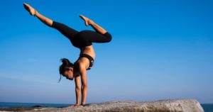 Promisecare Medical Group - A woman performing a handstand on a rock by the sea, her legs bent gracefully in the air against a clear blue sky.