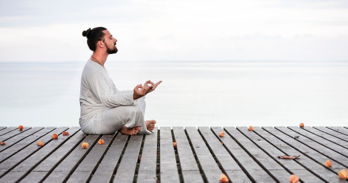 Promisecare Medical Group - A man in light clothing meditating in a lotus position on a wooden pier overlooking a calm sea, with flowers scattered around.