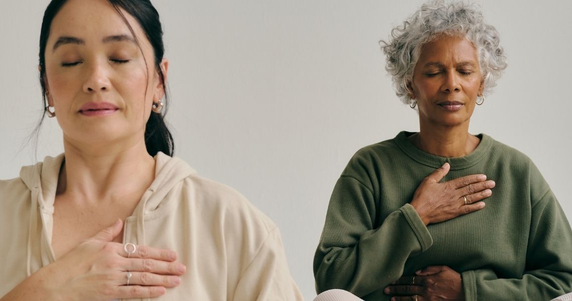 Promisecare Medical Group - Two women meditating, one young with long hair and the other elderly with short gray hair, both with eyes closed and hands on their chests.