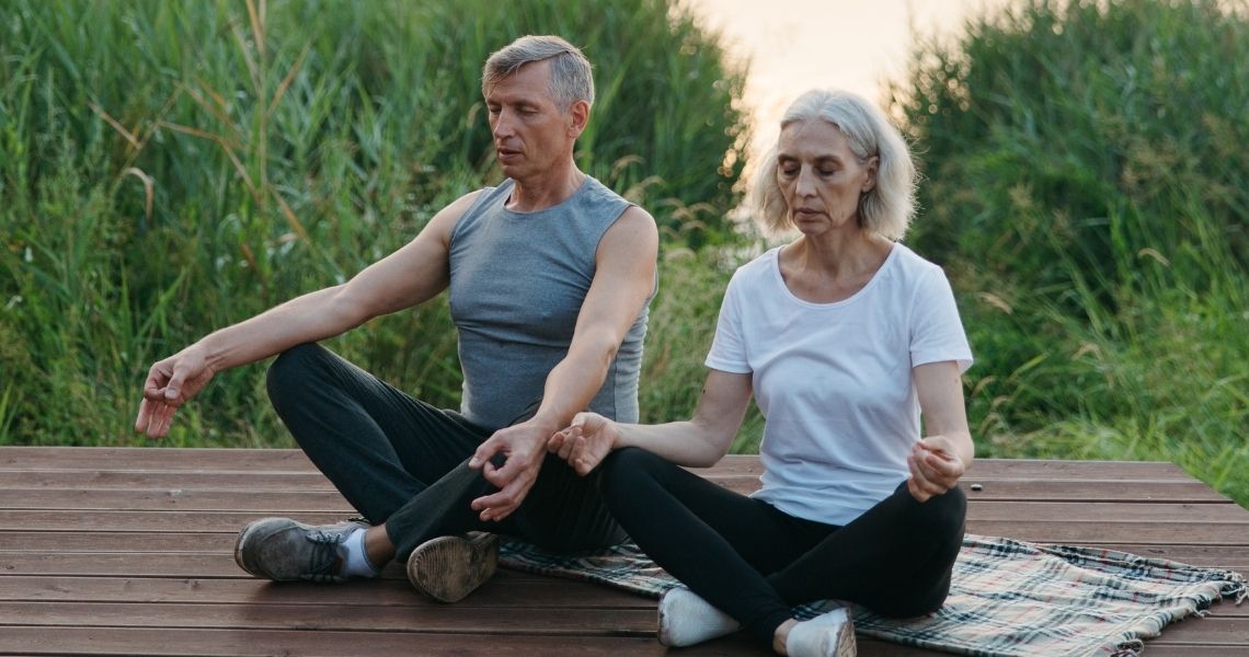 Promisecare Medical Group - An elderly man and woman meditating peacefully on a wooden deck surrounded by tall grass.