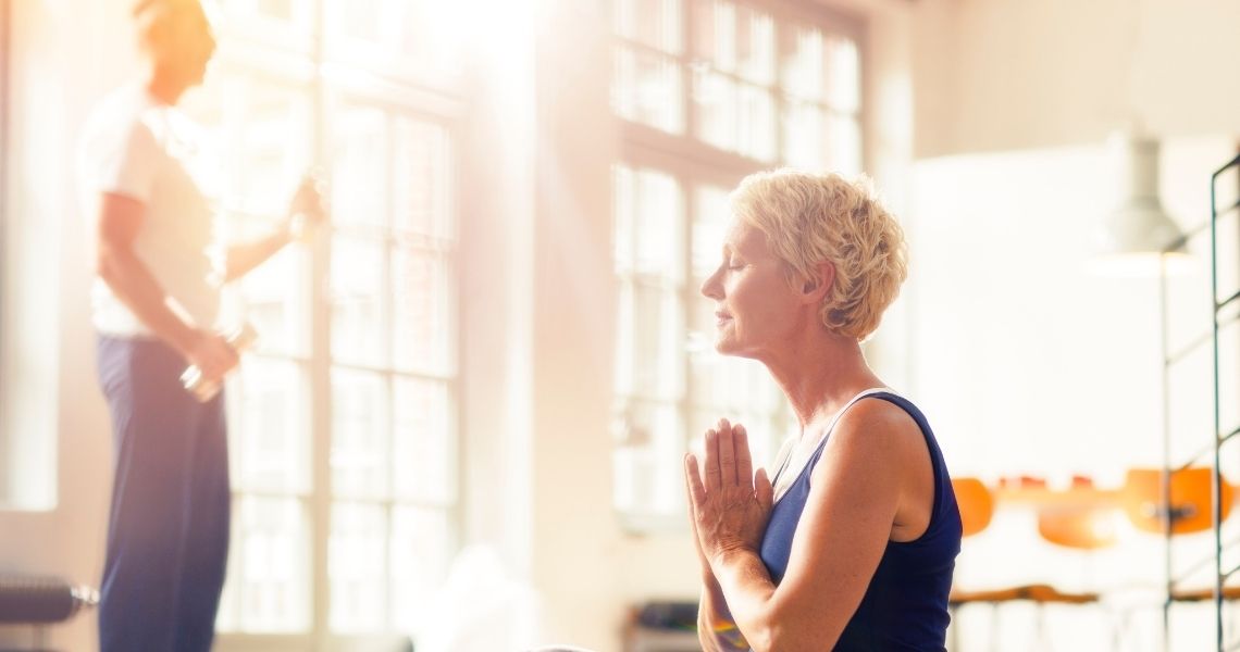 Promisecare Medical Group - An elderly woman practicing yoga in a sunlit room, with hands in prayer position, as her instructor guides the class in the background.