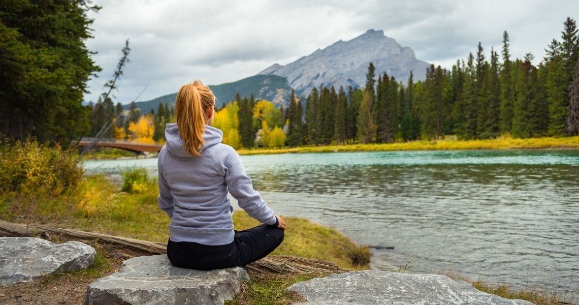 Promisecare Medical Group - A woman sitting cross-legged by a river, facing away from the camera, and looking towards a mountainous landscape with autumn-colored trees.