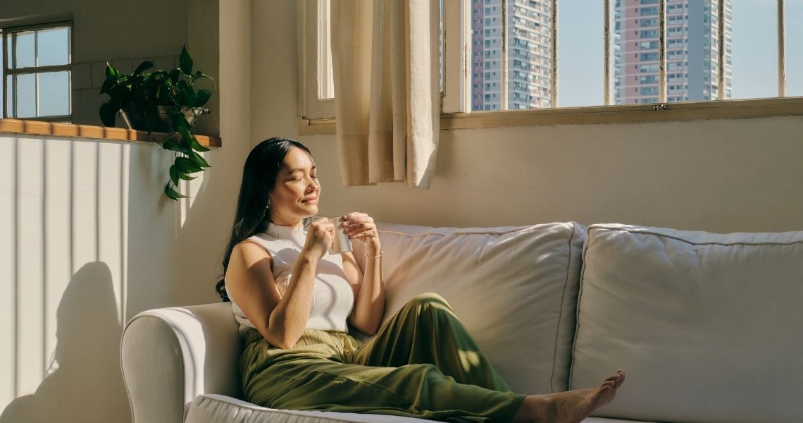 Promisecare Medical Group - A woman relaxing on a white sofa with a cup in her hands, bathed in sunlight, near a window overlooking high-rise buildings.